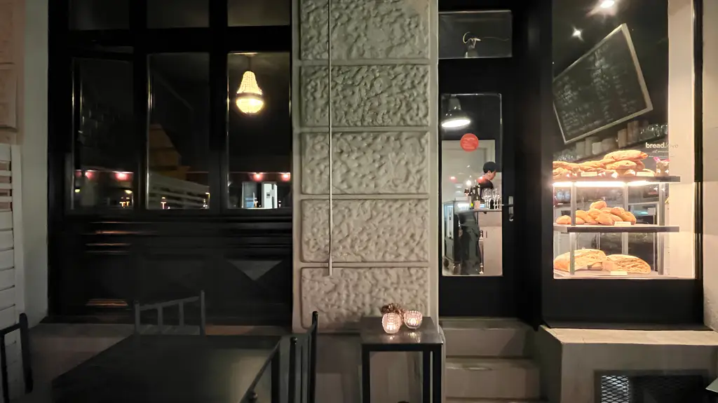 Abendliche Außenansicht eines Ladens mit beleuchteter Brotauslage, gläserner Tür, Kronleuchter und stilvollem Interieur durch großes Fenster sichtbar.
