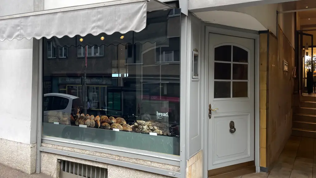 Blick auf Schaufenster mit Brotauslage und Eingang der Bäckerei bread.love mit grauer Tür und heller Markise.
