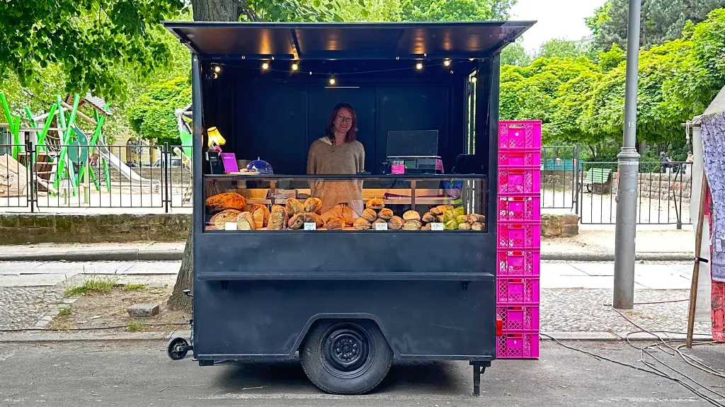bread.love Verkaufsstand in Berlin mit großer Brotauswahl, pinken Kisten und freundlicher Verkäuferin vor einem Spielplatz im Hintergrund.