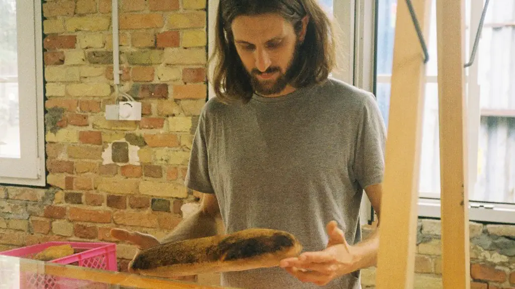 Mann mit langen Haaren und grauem T-Shirt hält ein längliches, dunkel gebackenes Brot vor einer Wand aus Backstein.