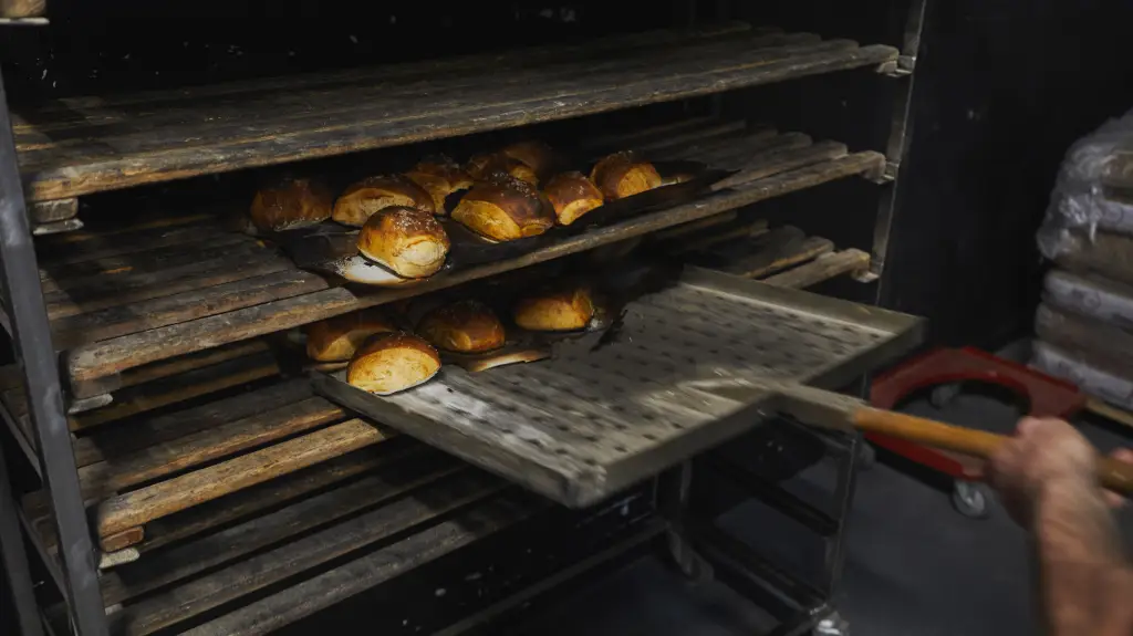 Ofenfrische Brötchen auf Backblechen werden mit einer flachen Schaufel in ein Regal mit Holzleisten einsortiert.
