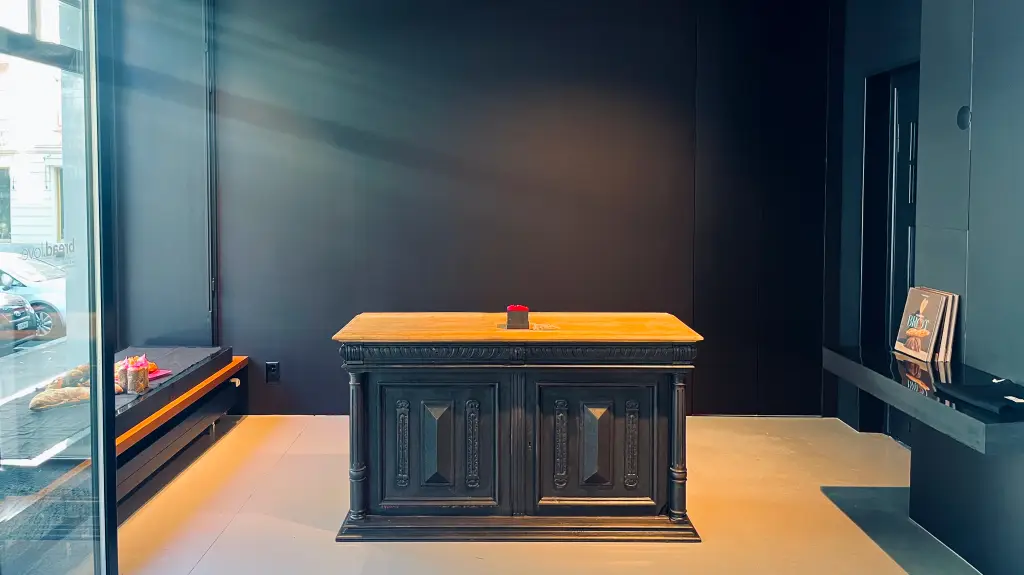 Innenraum mit massivem, dunkel verziertem Holztresen mit geschnitzten Paneelen vor schwarzer Wand, links im Fensterbereich Brotauslage.