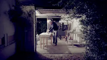Mann mit weißem Haar und dunkler Kleidung arbeitet nachts an einem Tisch in einer offenen Garage mit Backofen im Hintergrund.