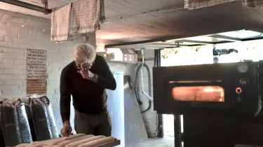 Mann mit weißem Haar und Brille prüft Teiglinge vor einem heißen Ofen in heller Garage mit Backofen, Warnschild und Tüchern an der Decke.