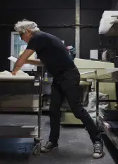 Mann mit weißem Haar, Brille und Stirnband bearbeitet Teiglinge an Edelstahltisch in Backstube mit dunklen Wänden und Geräten.