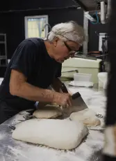 Mann mit weißem Haar, Stirnband und Brille teilt Teigstücke mit Teigschaber auf bemehlter Edelstahltisch in Backstube.
