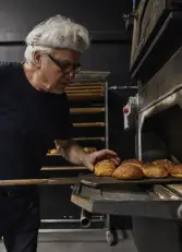 Mann mit weißem Haar, Brille und Stirnband nimmt mit Holzschieber frisch gebackene Brötchen aus einem Ofen in dunkler Backstube.