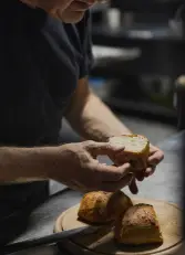 Person in dunklem T-Shirt hält ein aufgebrochenes Brötchen über Holzbrett mit drei knusprigen Brötchen in dunkler Backstube.