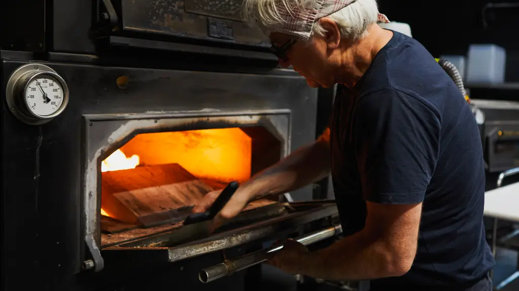 Mann mit weißem Haar, Brille und dunklem T-Shirt schiebt mit einer Holzschaufel Brot in einen heißen Ofen mit sichtbarer Glut.