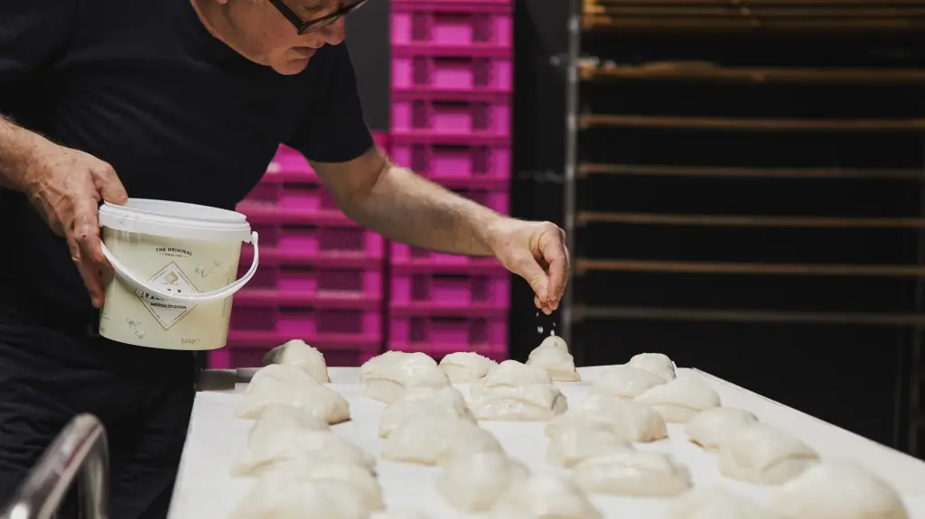 Mann mit schwarzem Shirt streut Salz auf Teiglinge auf Blech, in der Hand weißer Eimer, im Hintergrund pinke Kisten.